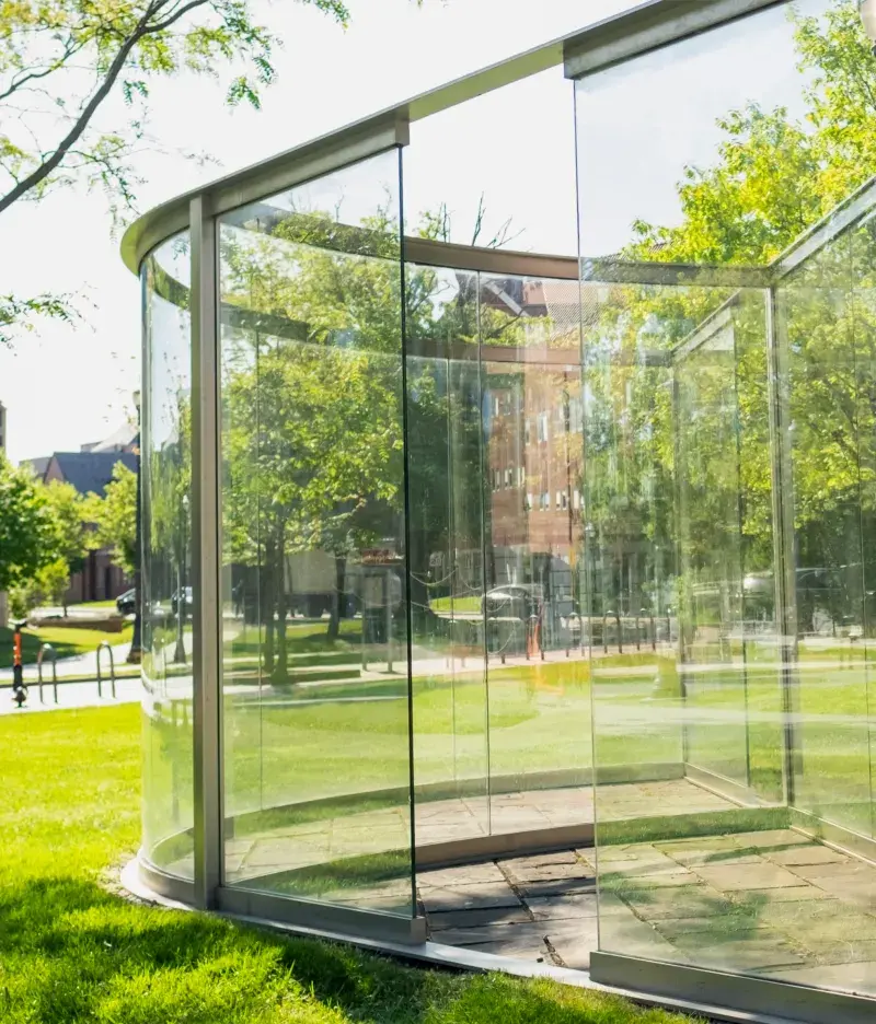 Large glass panes within a metal frame come to a point on one side and extend back, with one side curving. The sculpture sits in a grassy area.