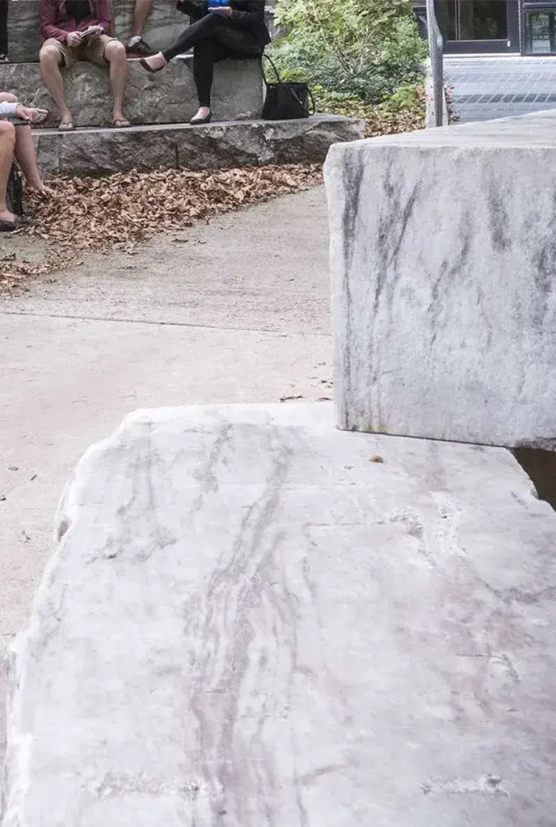 Close up of white and gray marble slabs sit alongside a walkway in a courtyard with trees. Students are sitting on more slabs in the background.