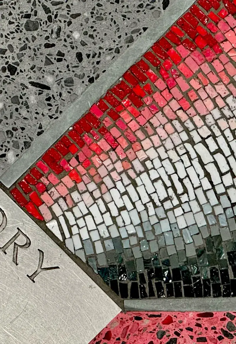 Close up of a circular floor installation with a red, gray, and black mosaic and a black and gray terrazzo pattern. 